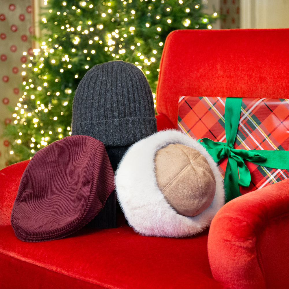 Three winter hats displayed on a red armchair with a blurred Christmas tree in the background: a men's charcoal grey ribbed knit beanie hat, a men's burgundy corduroy flat cap, and a women's white faux-fur hat with a suede top. A wrapped holiday gift sits beside them.