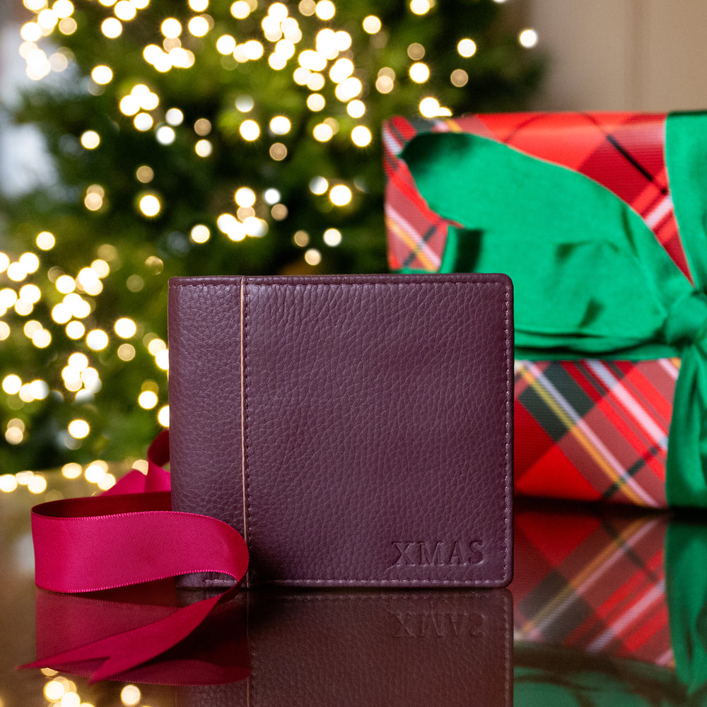 A leather bifold wallet with 'XMAS' embossed on the front sat on a table with a red ribbon and a Christmas gift box in the background against a blurred Christmas tree.