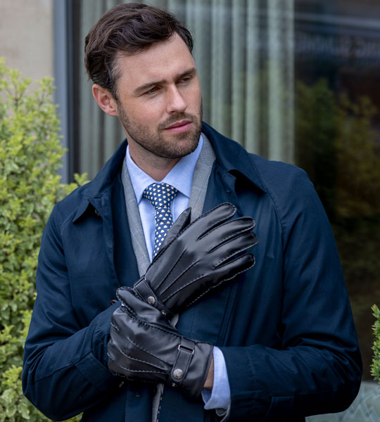 A man wearing black leather gloves, standing outdoors in front of a building with greenery.
