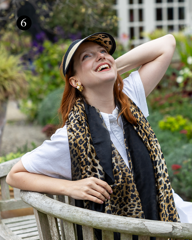 Woman smiling in garden wearing animal print scarf and matching visor, sitting on a bench.
