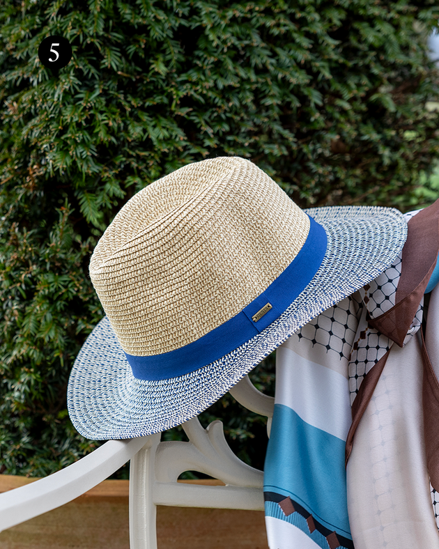 Women's straw fedora hat with two-colour brim and tonal colour band in navy hanging on a chair in the garden.